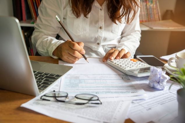 Imagem de uma mulher fazendo cálculos, com um laptop na mesa, calculadora e papéis