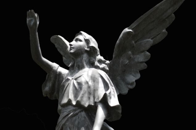 Imagem da estátua de um anjo em um fundo preto com as mãos e cabeça erguidas pra cima e seu rosto parece sereno