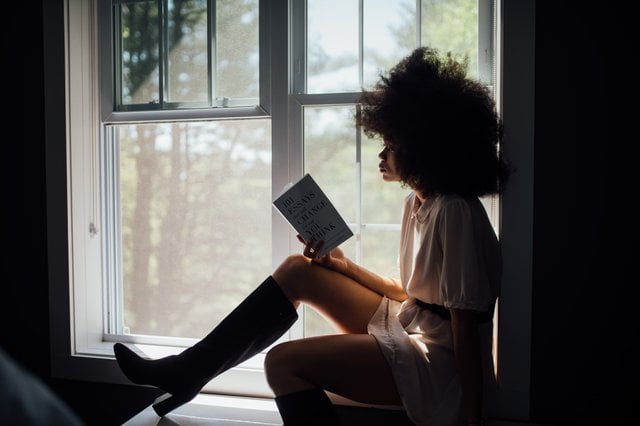 Mulher negra sentada numa janela lendo um livro.