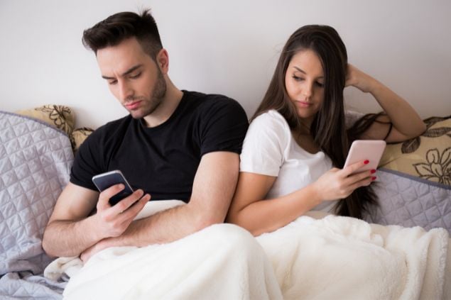 Imagem de um casal sentado na cama e olhando o celular, a mulher está tentando ver o celular de seu parceiro