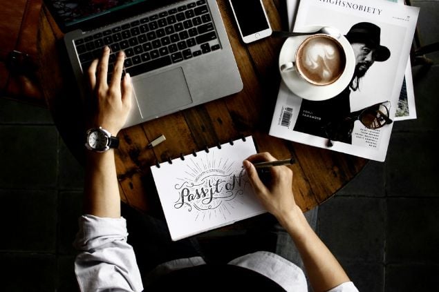 Imagem vista de cima, de uma pessoa fazendo planejamentos em sua mesa de escritório, na frente do laptop e escrevendo e desenhando em um caderno