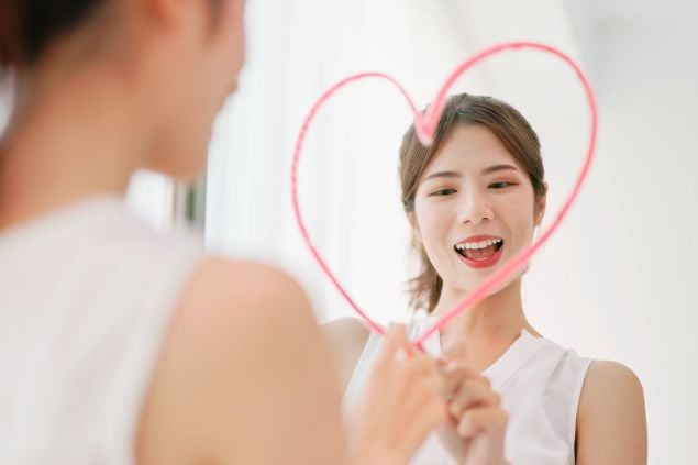Imagem de uma mulher na frente do espelho sorrindo e desenhando um coração no espelho