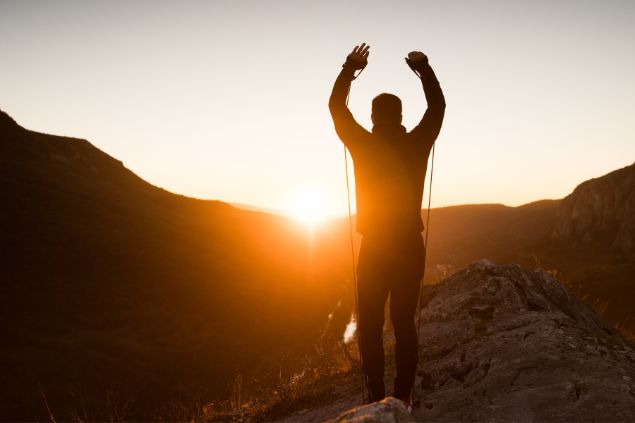 Imagem de uma pessoa no topo de uma montanha de frente para o por do sol com os braços levantados