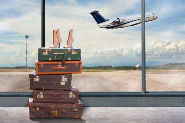 Imagem da janela de aeroporto, malas empilhadas em frente dela e um sapato em cima. E do lado de fora a vista de um avião decolando
