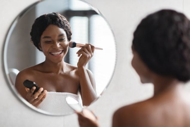 Imagem de uma mulher negra se maquiando no espelho com um sorriso largo