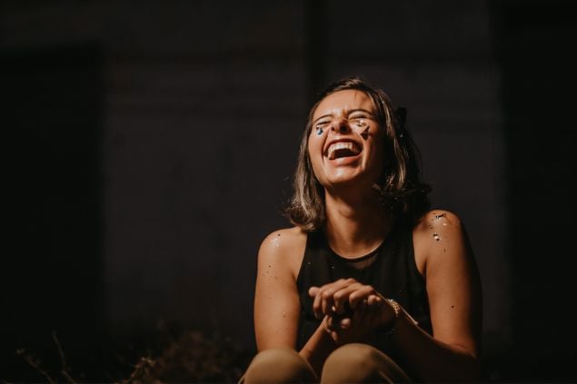 Imagem de uma mulher branca com a cabeça erguida, os olhos fechados e dando risada em um fundo preto
