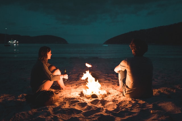 Homem e mulher sentados na beira da praia, ao redor de uma fogueira